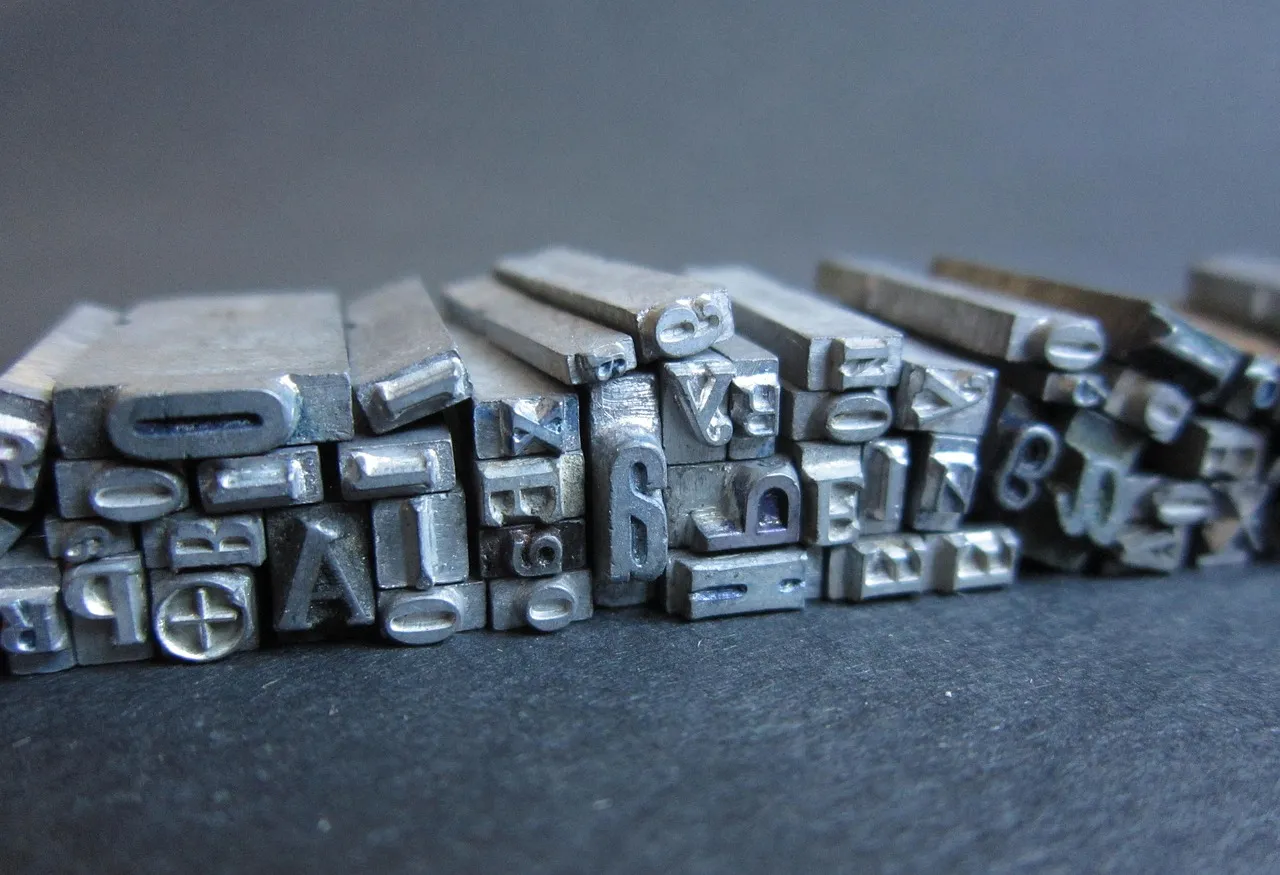 A close-up row of metal movable type letters standing on a dark surface, letterforms reversed and worn