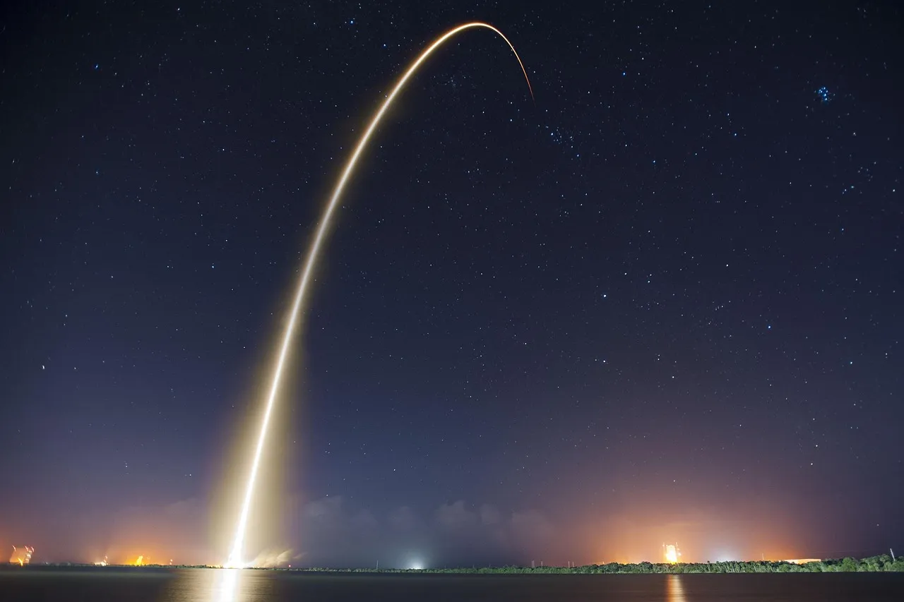 A long-exposure photograph of a rocket launch at night, its exhaust drawing a bright curved arc across a starfield above a coastline