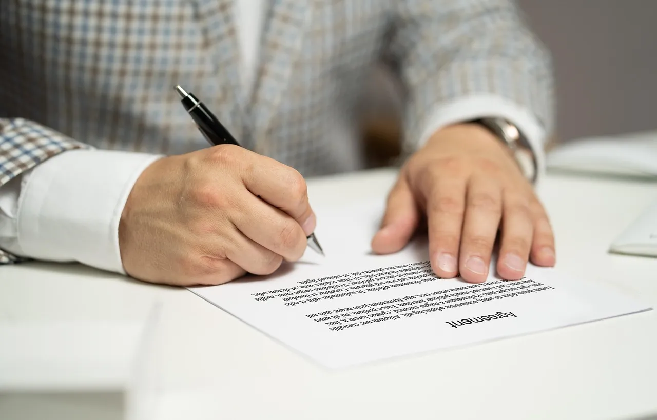 A person signing a printed agreement at a desk with a fountain pen.