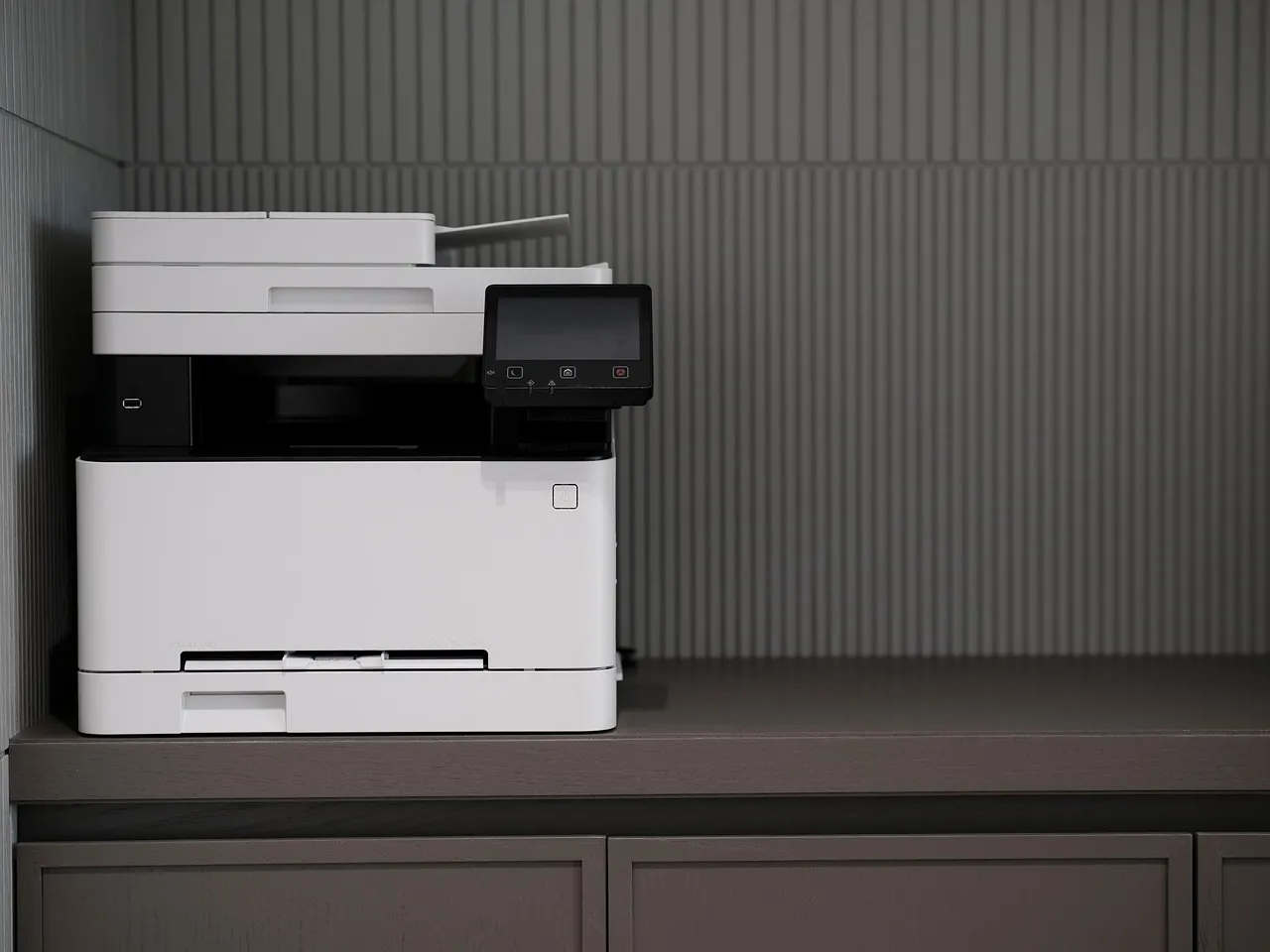A white office multifunction printer and scanner standing against a grey wall.