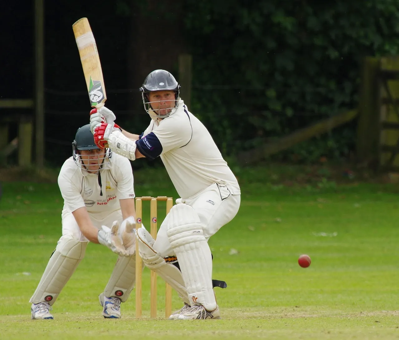A batsman in whites playing a shot with the wicketkeeper crouched behind the stumps and the red ball in the air