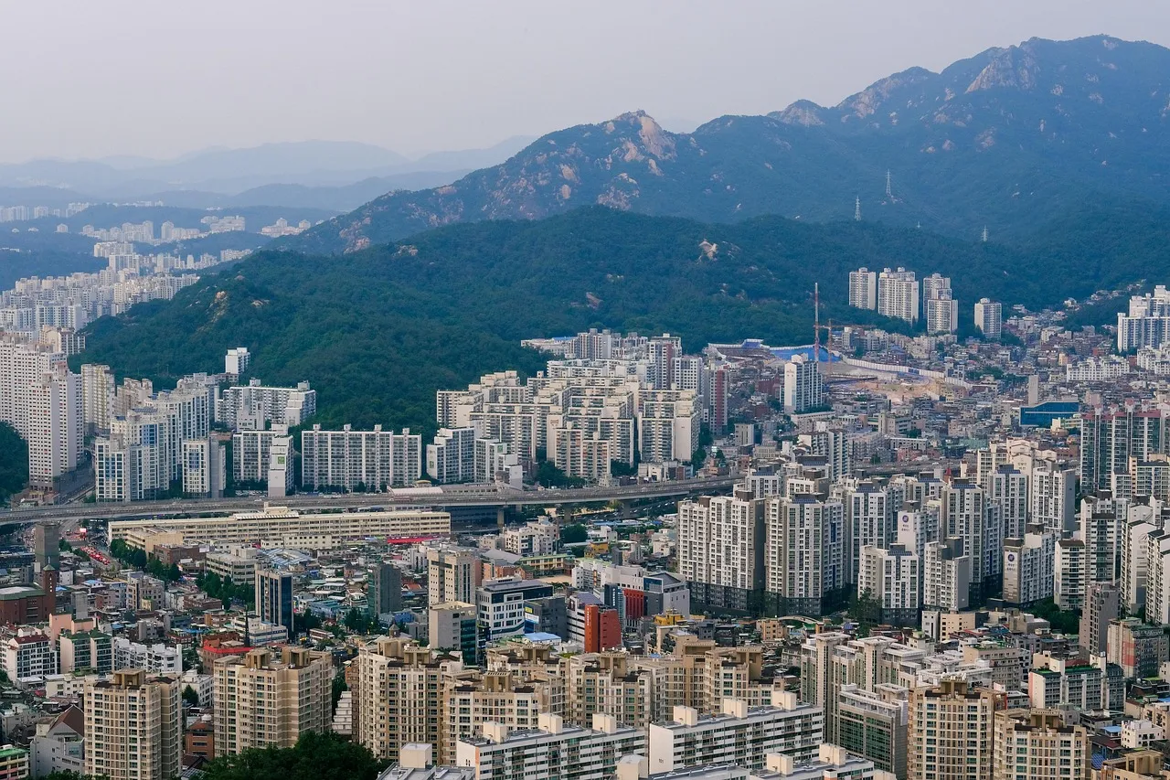 A wide view across Seoul's apartment blocks and office towers, with forested mountains behind.