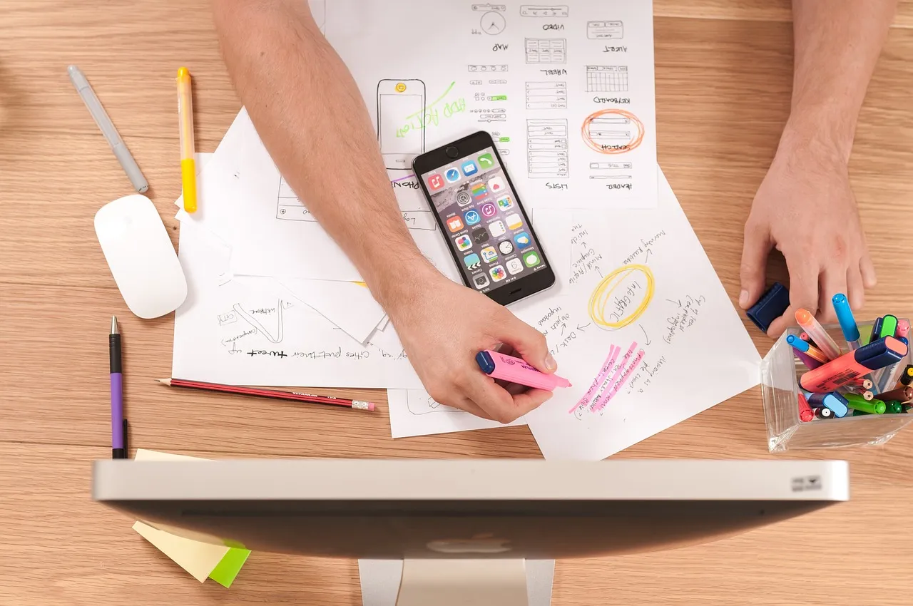 Overhead view of hands sketching app screen layouts on paper beside a smartphone, pens and a mouse.