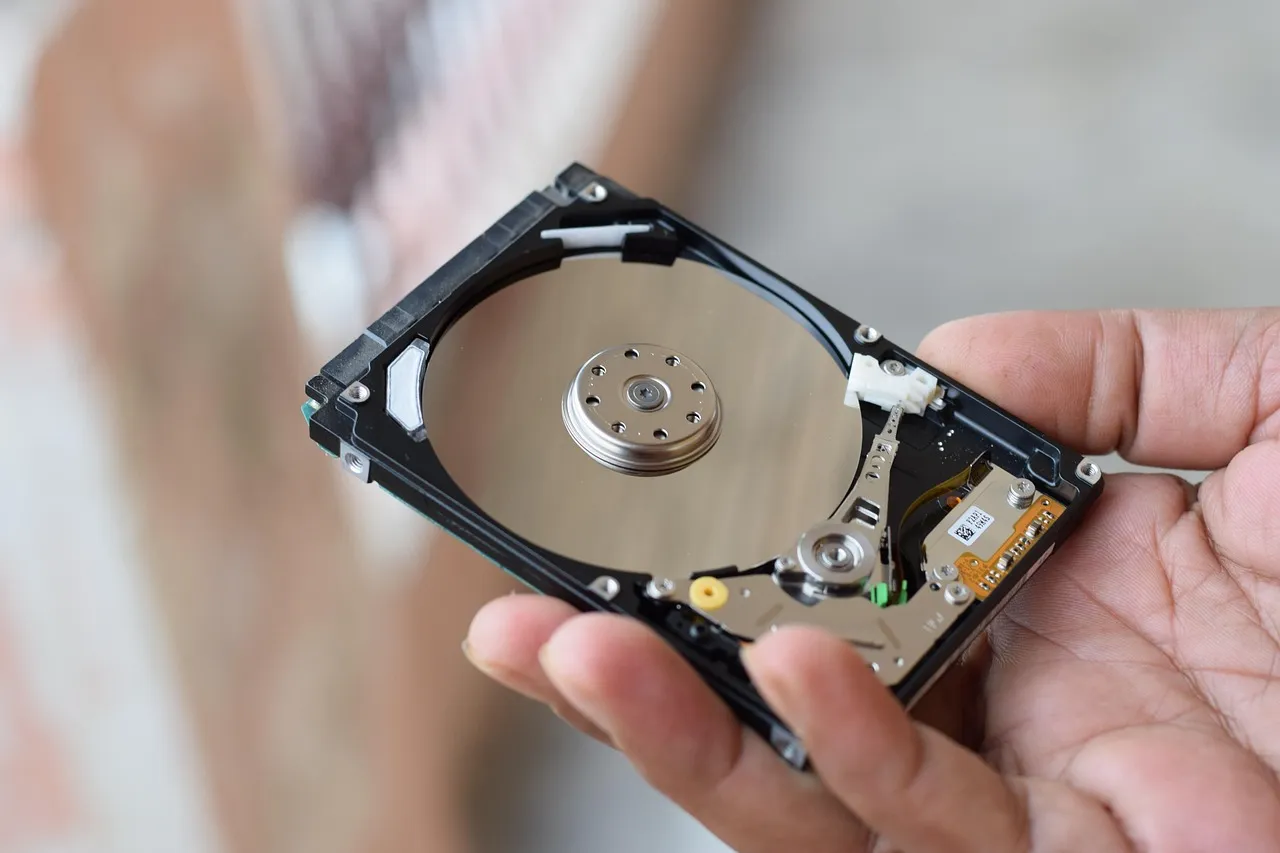 A hand holding an opened mechanical hard drive with its platter and read head exposed.