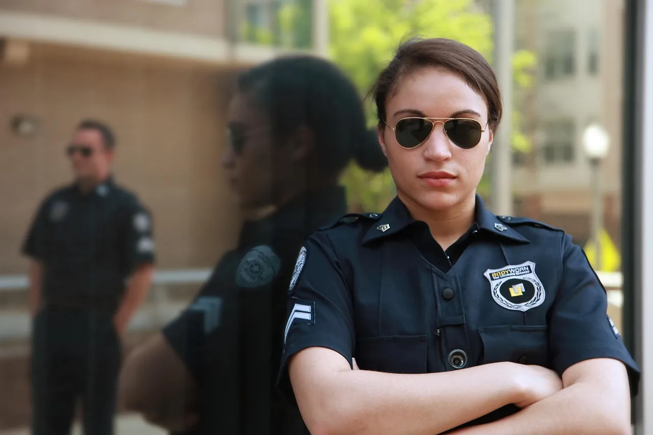 A uniformed police officer in sunglasses stands with arms folded outside a building, with a second officer out of focus behind