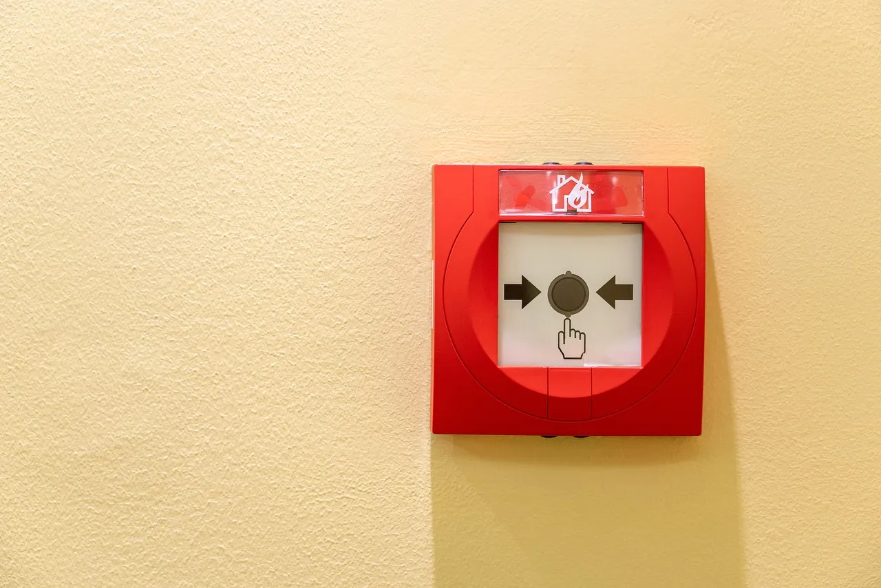 A red emergency call point mounted on a plain wall, its pictogram showing a hand pressing the central button.