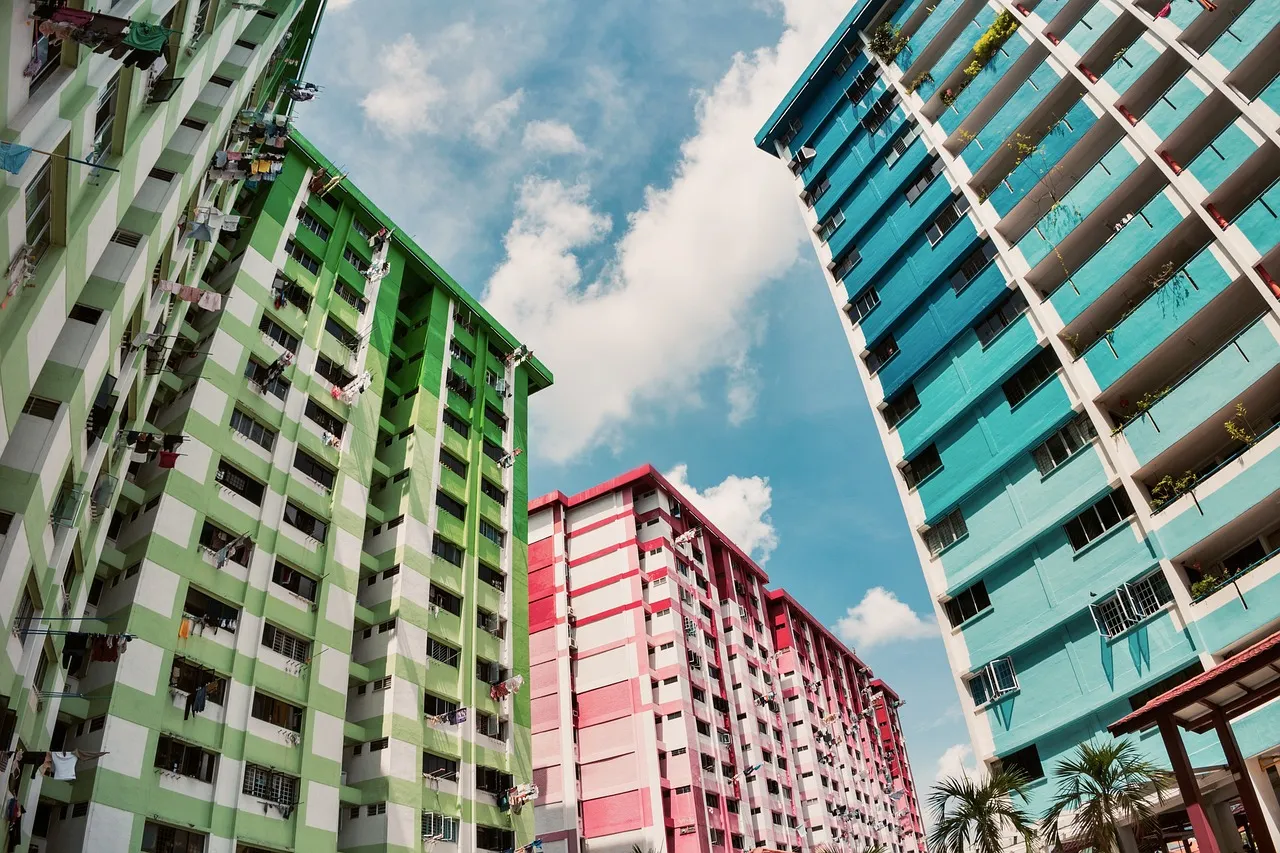 Three Singapore HDB blocks in green, pink and teal seen from below, laundry poles projecting from the flats against a blue sky.
