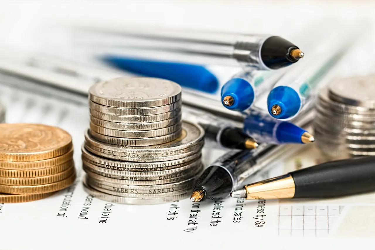 Stacks of coins of unequal height resting on a printed financial document, with several ballpoint pens laid across it