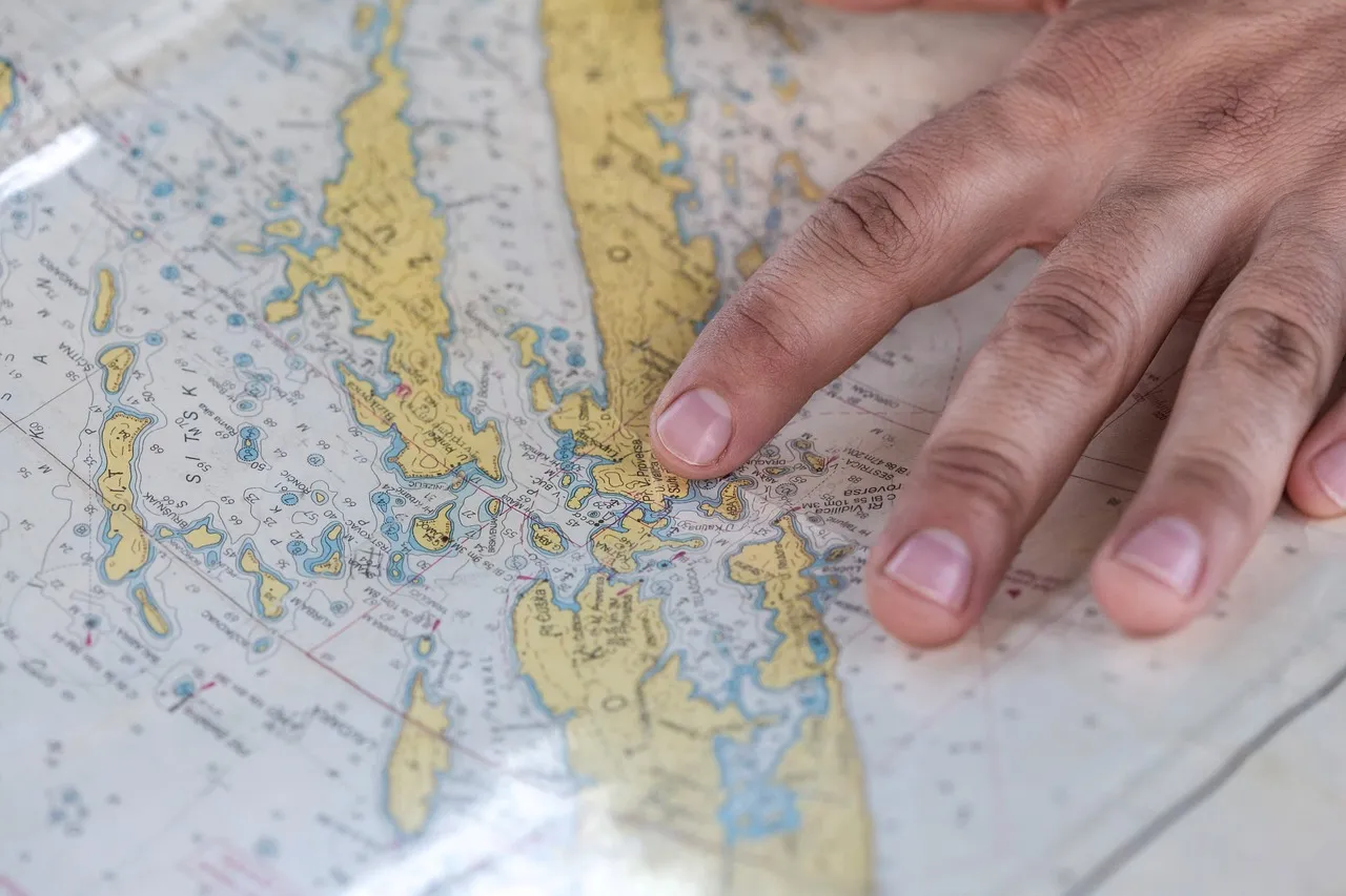 A hand resting on a printed nautical chart, one finger marking a position among islands and depth soundings