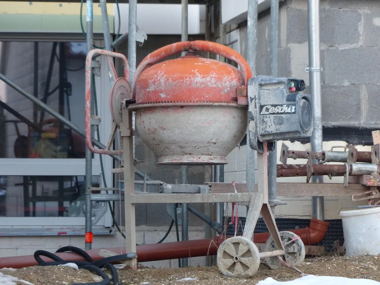 A well-used orange and grey electric cement mixer standing on a building site, its drum crusted with dried mortar, beside scaffolding poles and a plastic bucket.