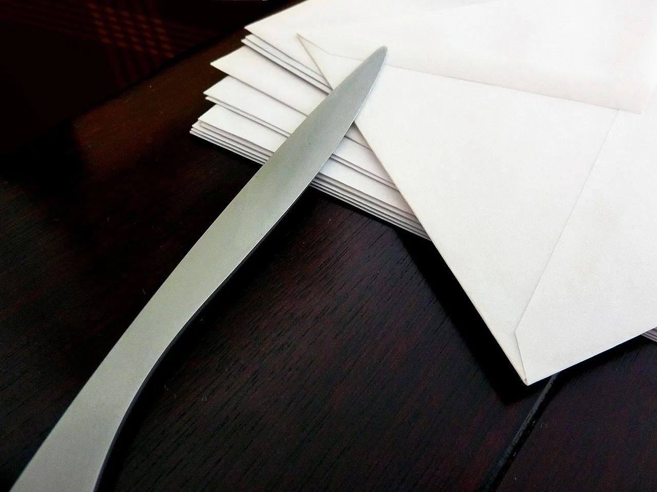 A stack of sealed white envelopes on a dark wooden desk with a metal letter opener resting across them