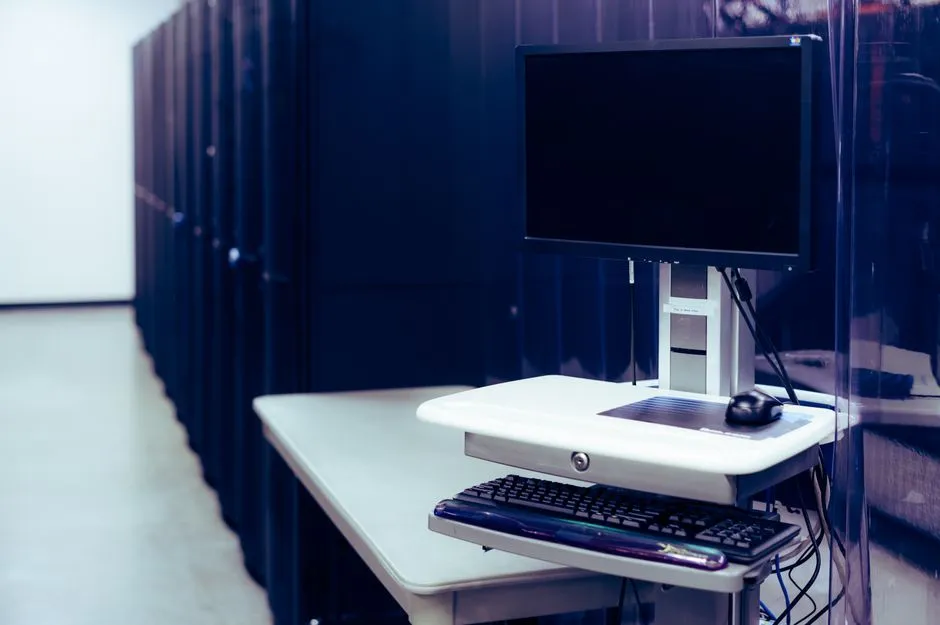 A computer with a black screen on a stand beside a row of steel server racks in a data centre.